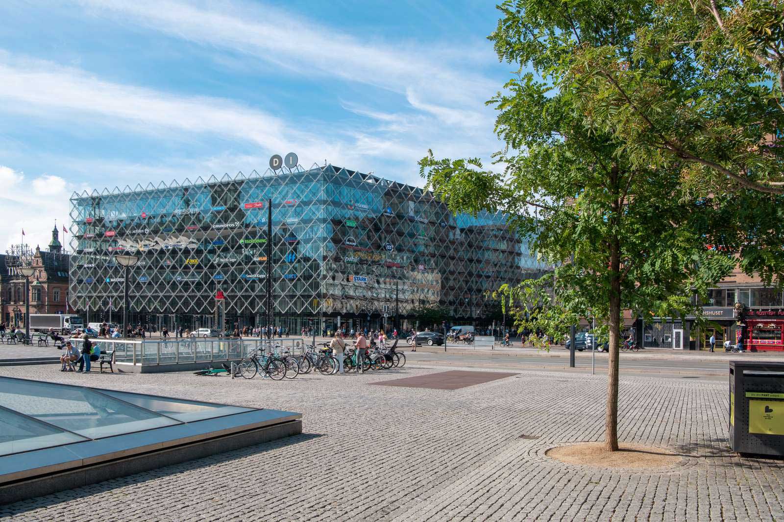 A modern glass building with various brand logos, surrounded by trees and bustling streets filled with pedestrians and bicycles.
