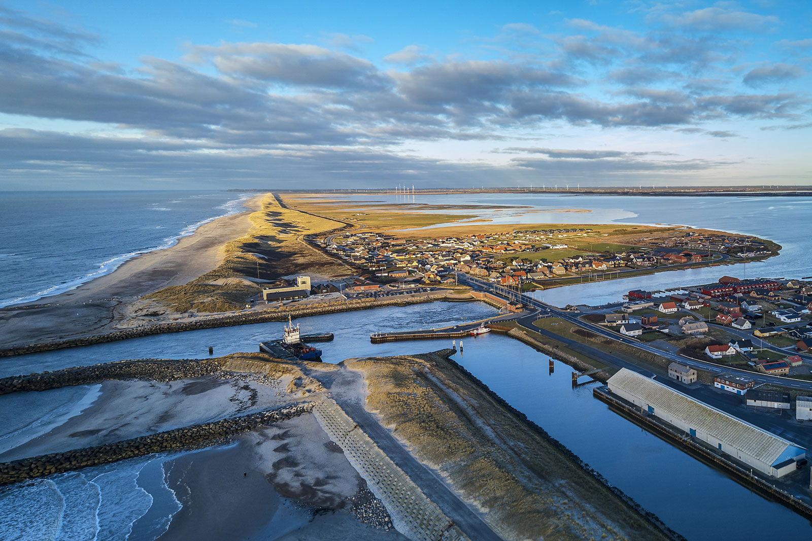 Aerial view of a coastal village with sandy beaches, a winding river, and wind turbines visible on the horizon.
