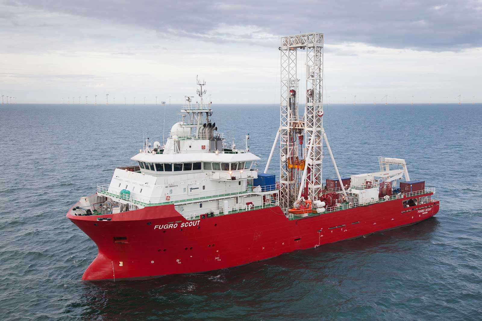 A red research vessel named 'FUGRO SCOUT' is seen in the ocean with drilling equipment on board. Wind turbines are visible in the background.