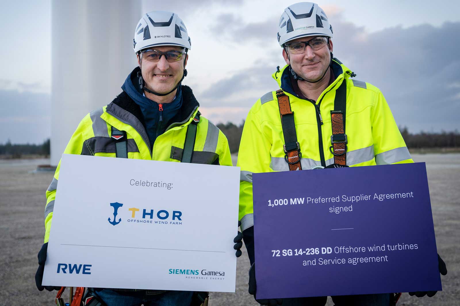 Two workers in high-visibility jackets celebrate the THOR Offshore Wind Farm agreement, holding signs with details about the project.