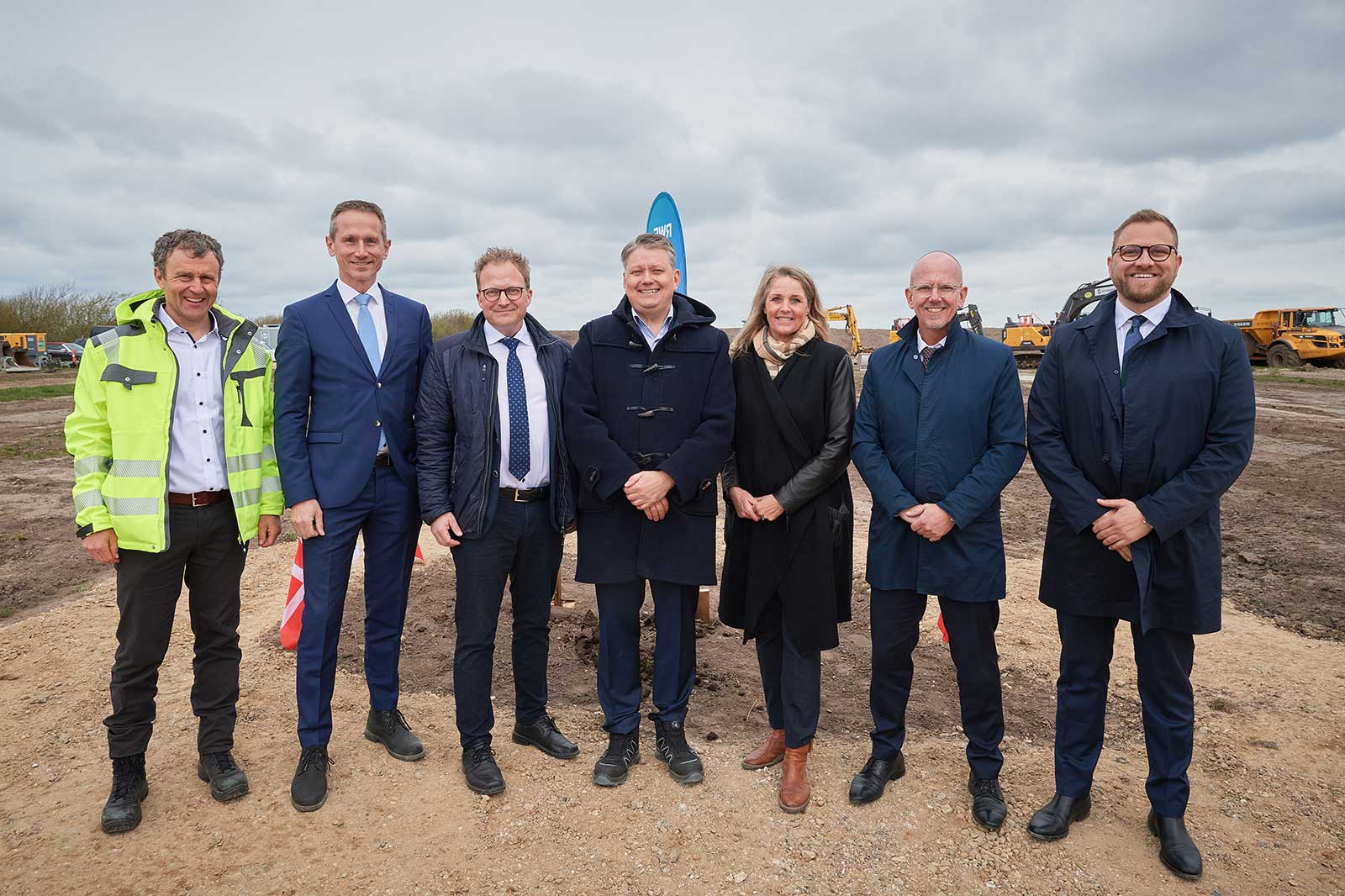 A group of professionals stands together at a construction site, with machinery in the background and an overcast sky.