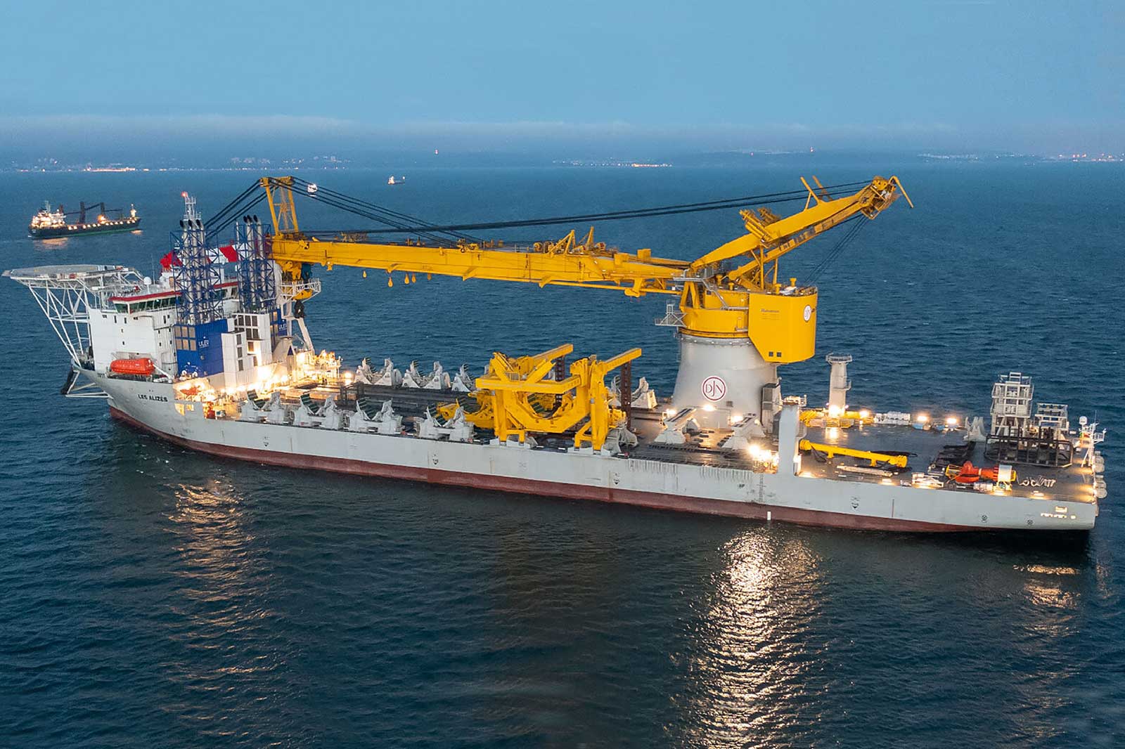 A large ship with a prominent yellow crane is positioned on the water, illuminated by lights, with cargo containers on deck.