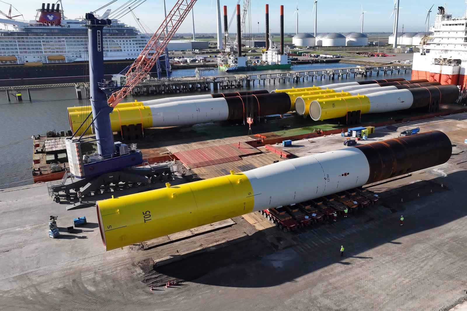 An aerial view of large yellow and white cylindrical structures at a dock, with a crane and a ship in the background.