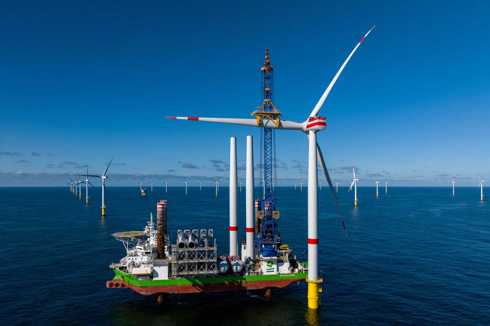 A large offshore wind turbine installation vessel next to a wind turbine, with multiple wind turbines in the background against a clear blue sky.
