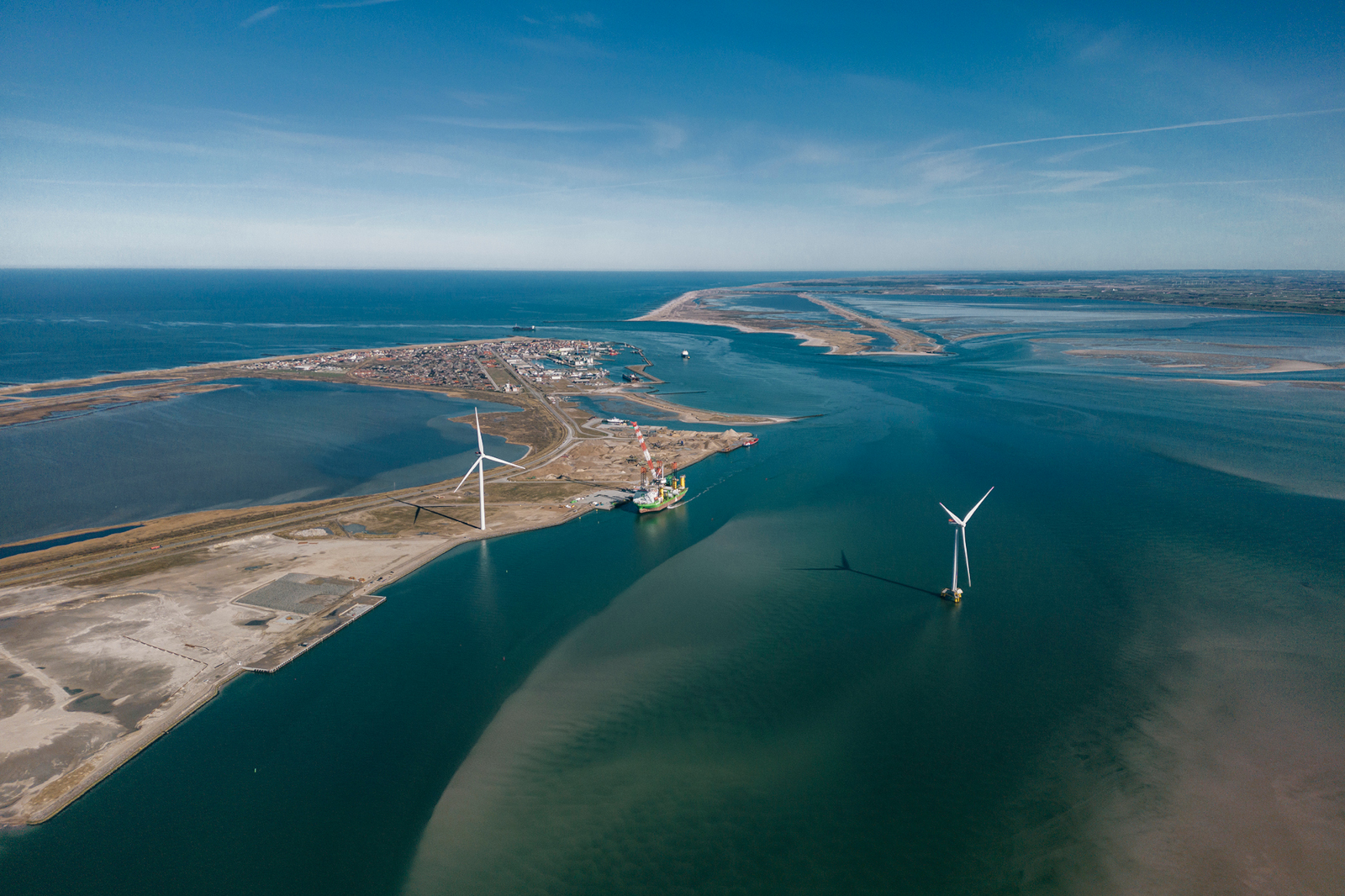 An aerial view of a coastal area featuring wind turbines, calm waters, and a small settlement in the distance under a clear sky.