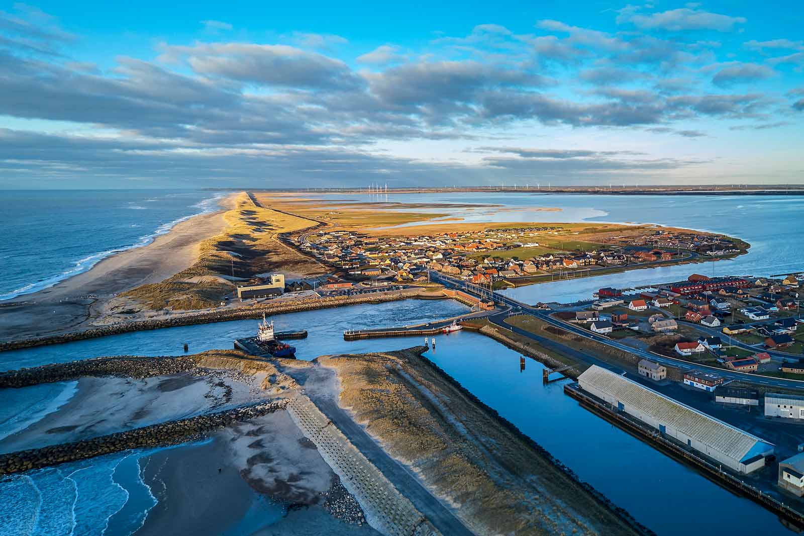 Et luftfoto af en kystby med strand, både og vand. Landskabet omfatter grønne marker og små huse.