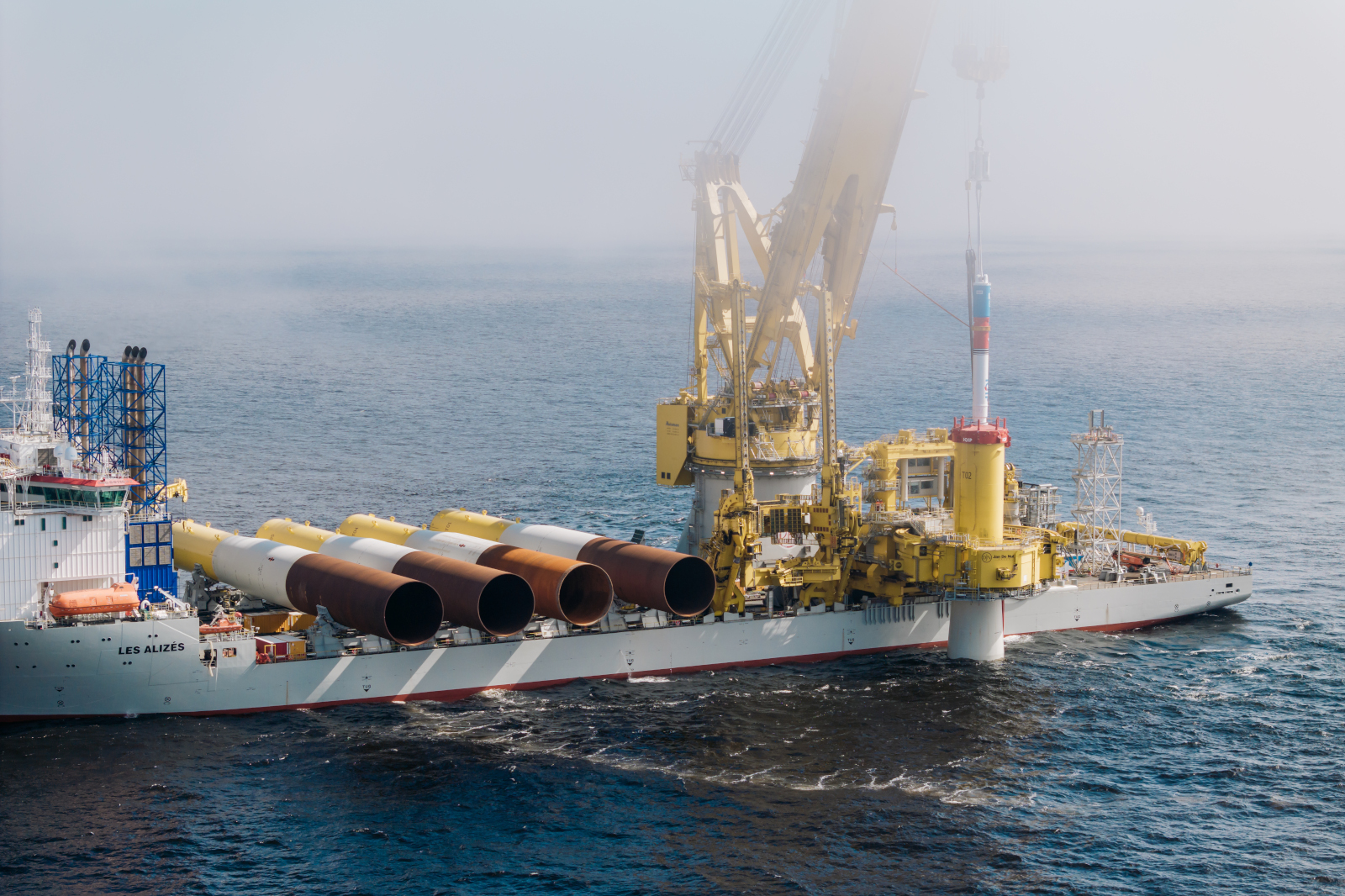 An offshore vessel, Les Alizés, operates with large pipes in the sea under a foggy sky.