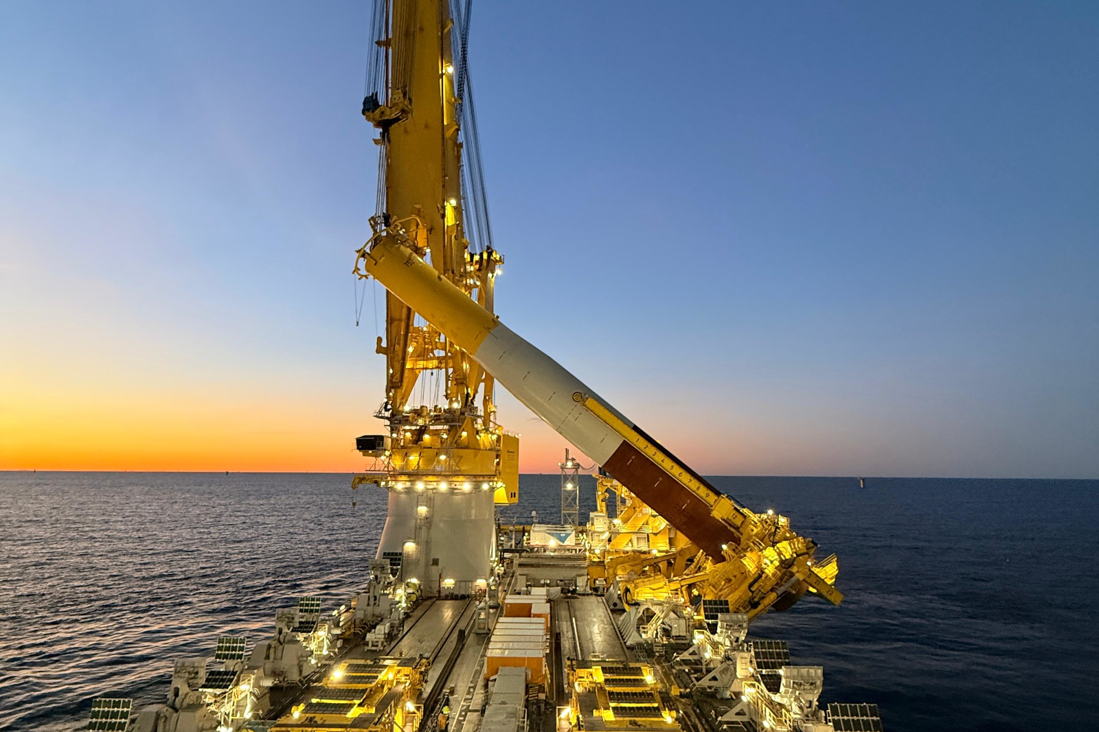 A large vessel with a yellow crane extending towards the horizon over the ocean during a colourful sunset.