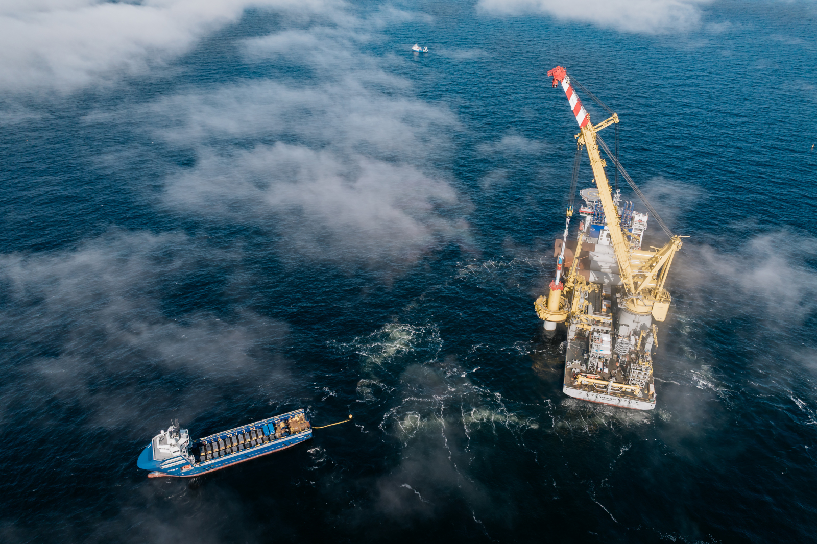 Et offshore boreplatform med en kran til højre og et skib i forgrunden, der arbejder i det blå hav med tåge.