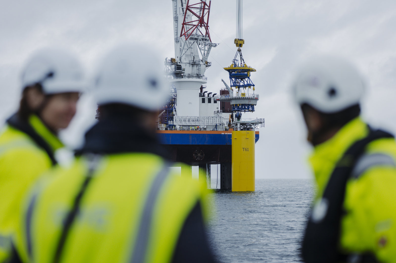 Three people in helmets and yellow jackets watch an offshore oil platform under a grey sky.