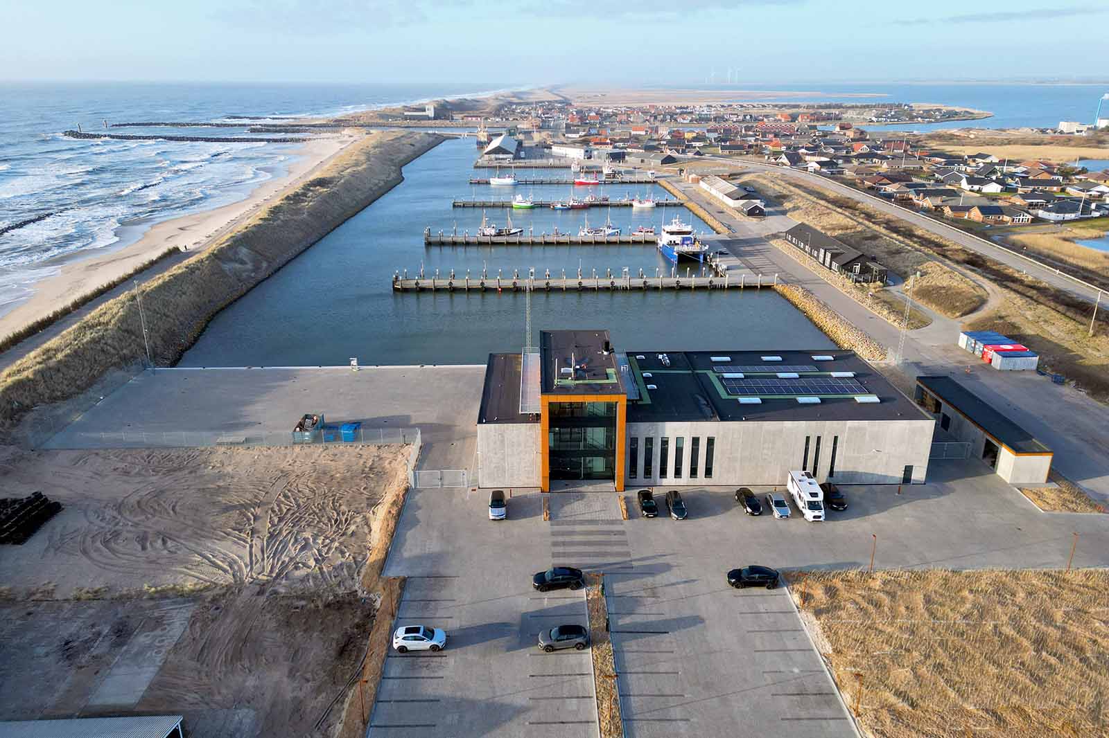 A modern building near a marina with several boats, surrounded by a coastal town, sandy beaches, and open sea in the background.