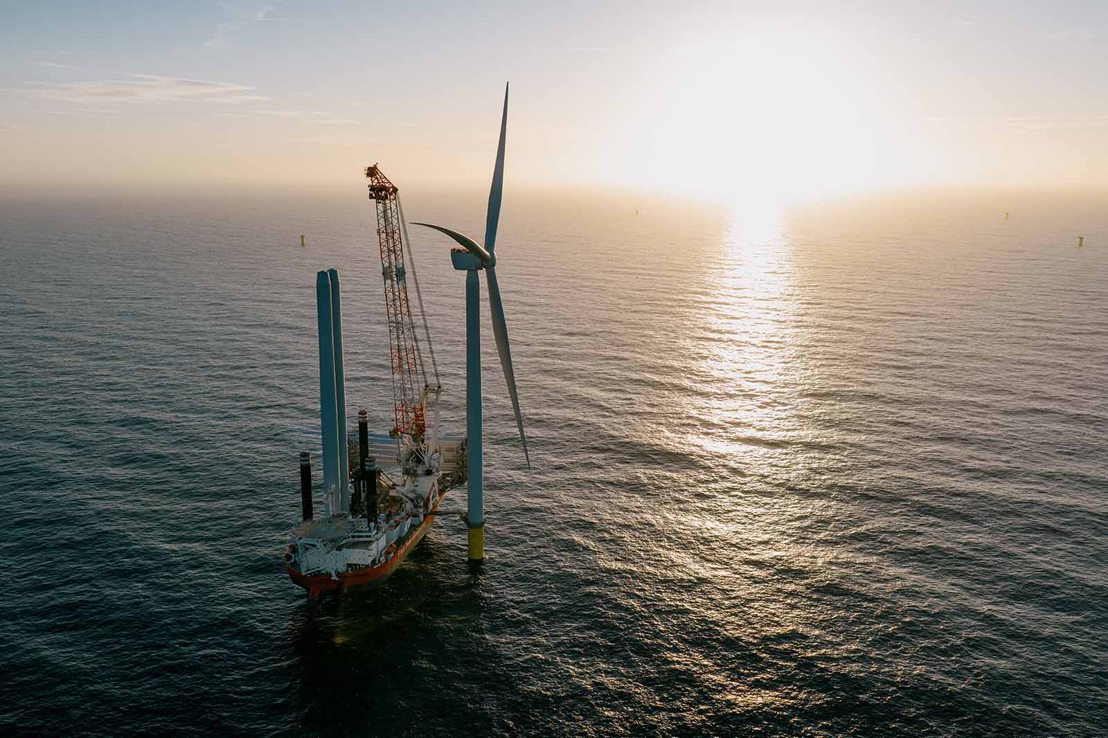 A vessel with a crane is working on an offshore wind turbine at sunset, with the ocean surface reflecting sunlight.