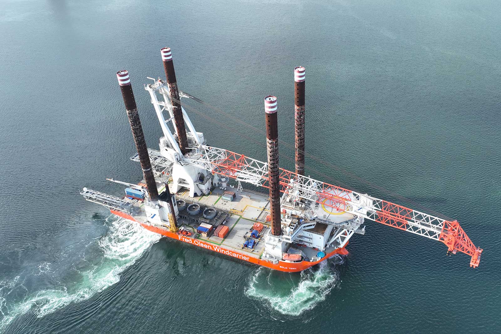 An aerial view of the Fred Olsen Windcarrier ship with three tall towers, navigating in calm waters.