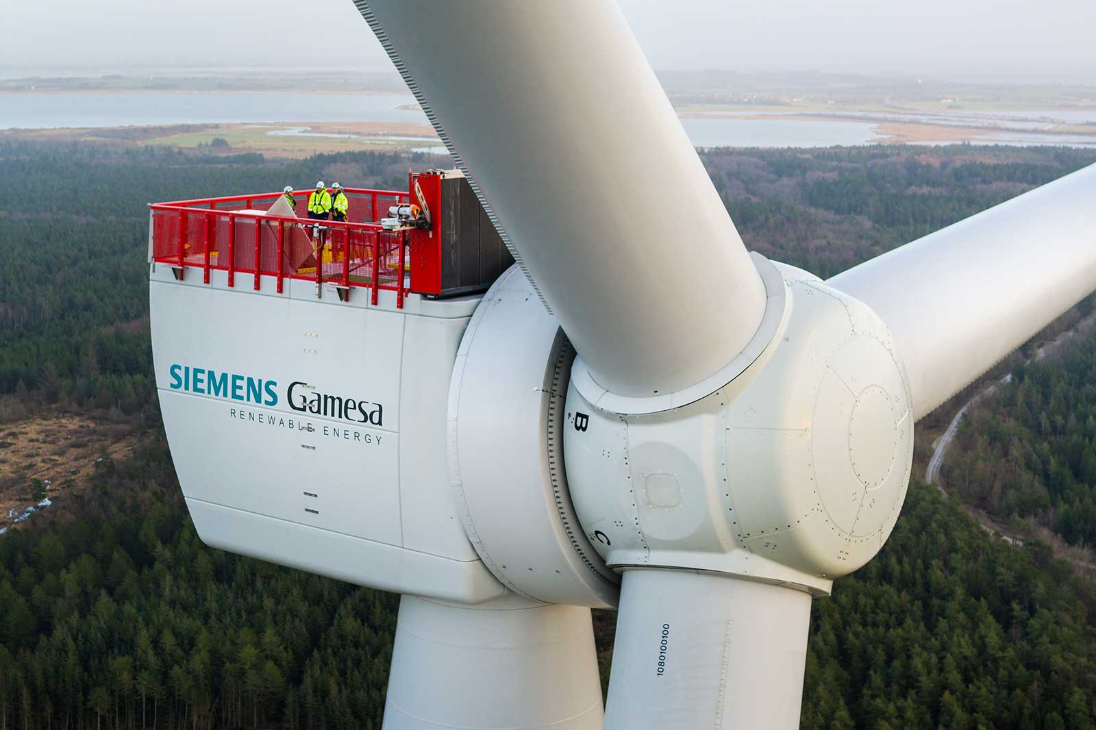 A Siemens Gamesa wind turbine with technicians on the platform, overlooking a lush forested area and water in the distance.