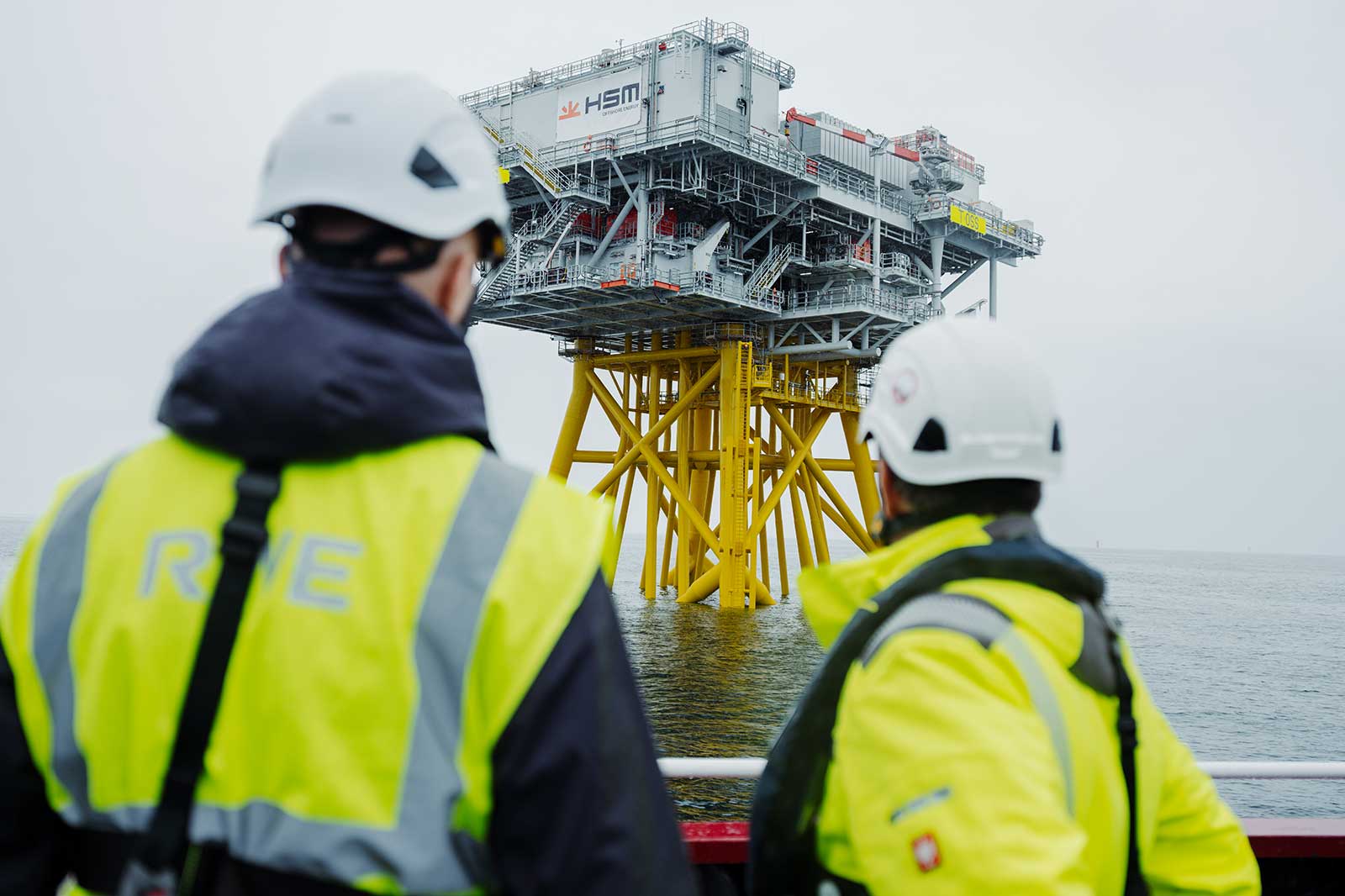 Two individuals in hard hats observe an offshore platform in the North Sea. The platform rests on yellow legs.