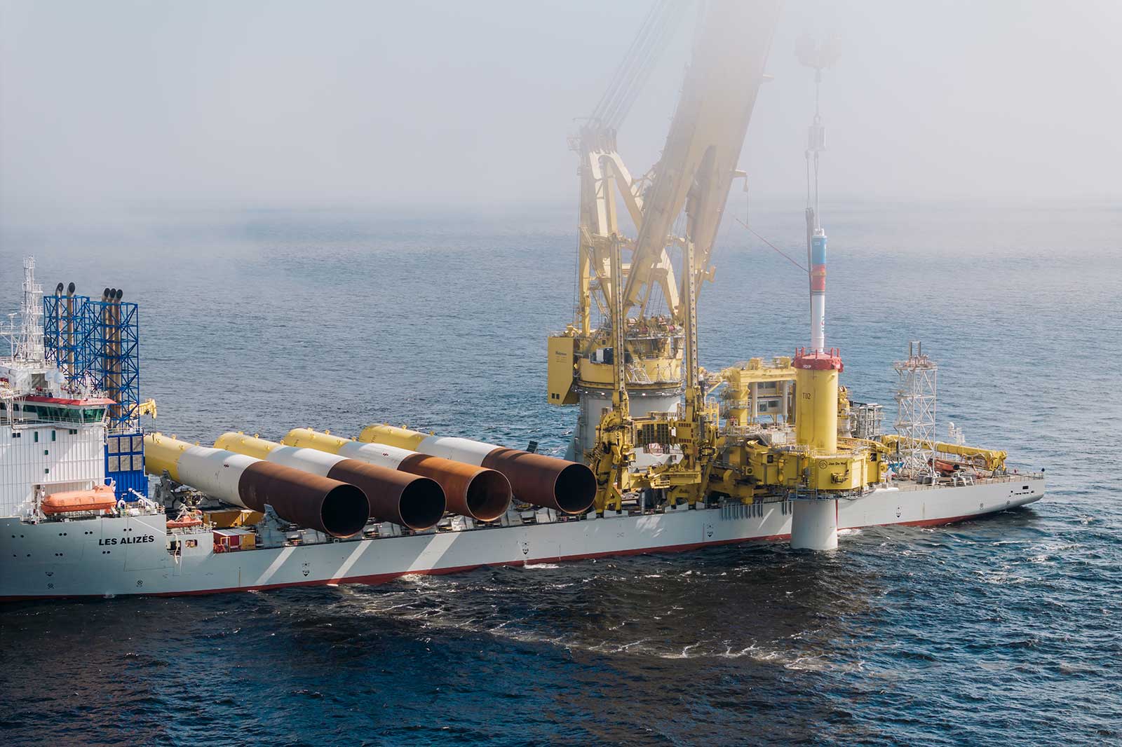 A large vessel with a yellow crane lifts large tubes over the sea in foggy weather.