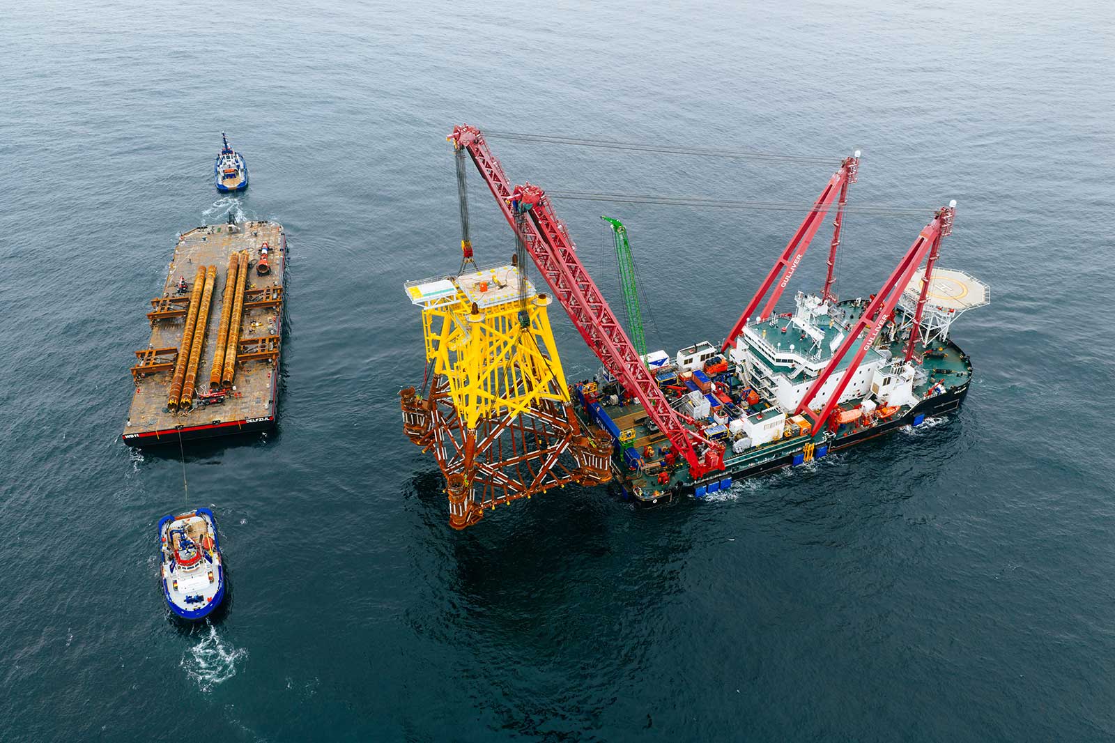 A large vessel with red cranes lifts a yellow offshore platform above water. Two smaller boats navigate nearby.