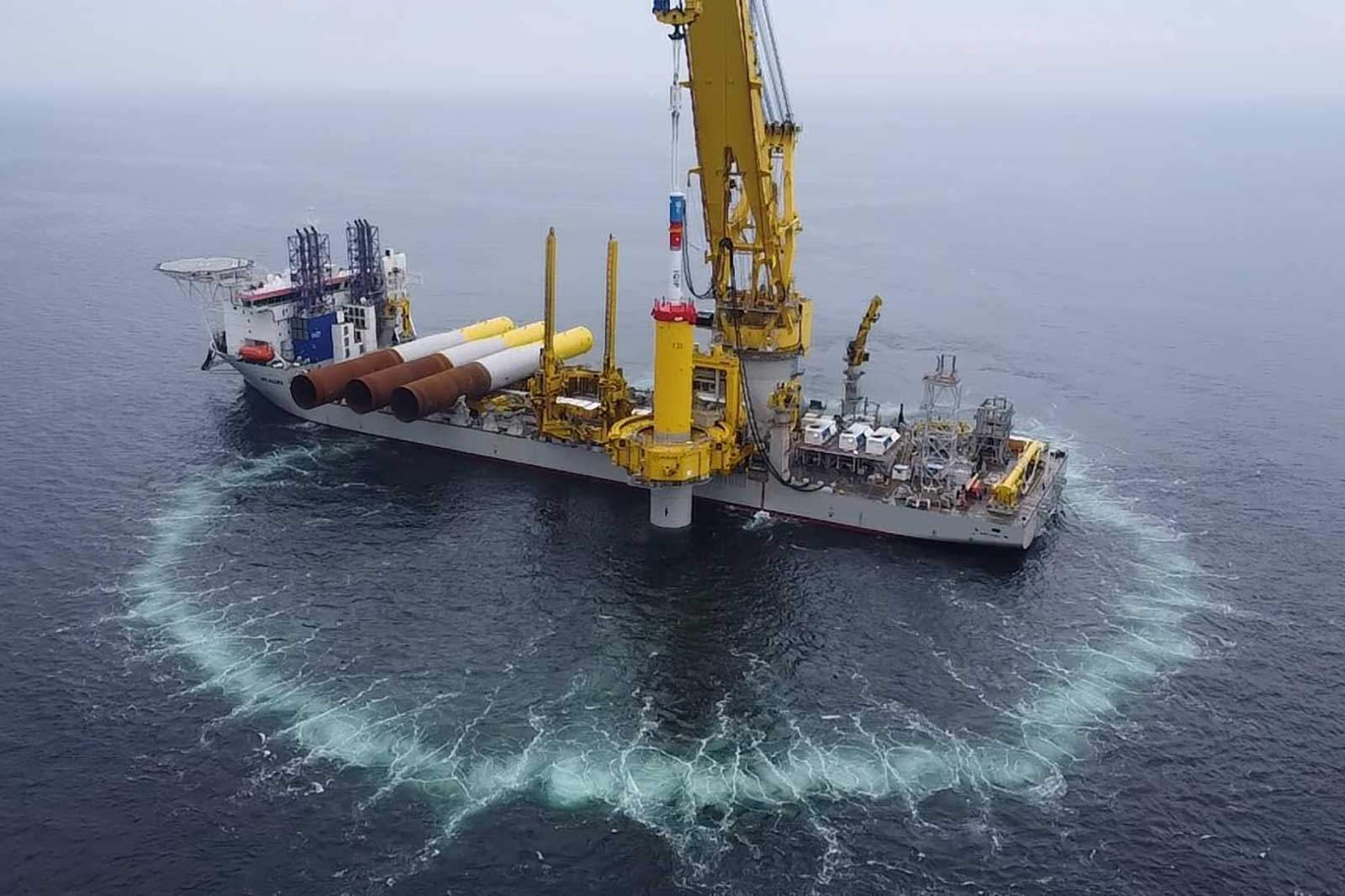 A ship with cranes and pipes works at sea, surrounded by circular waves.