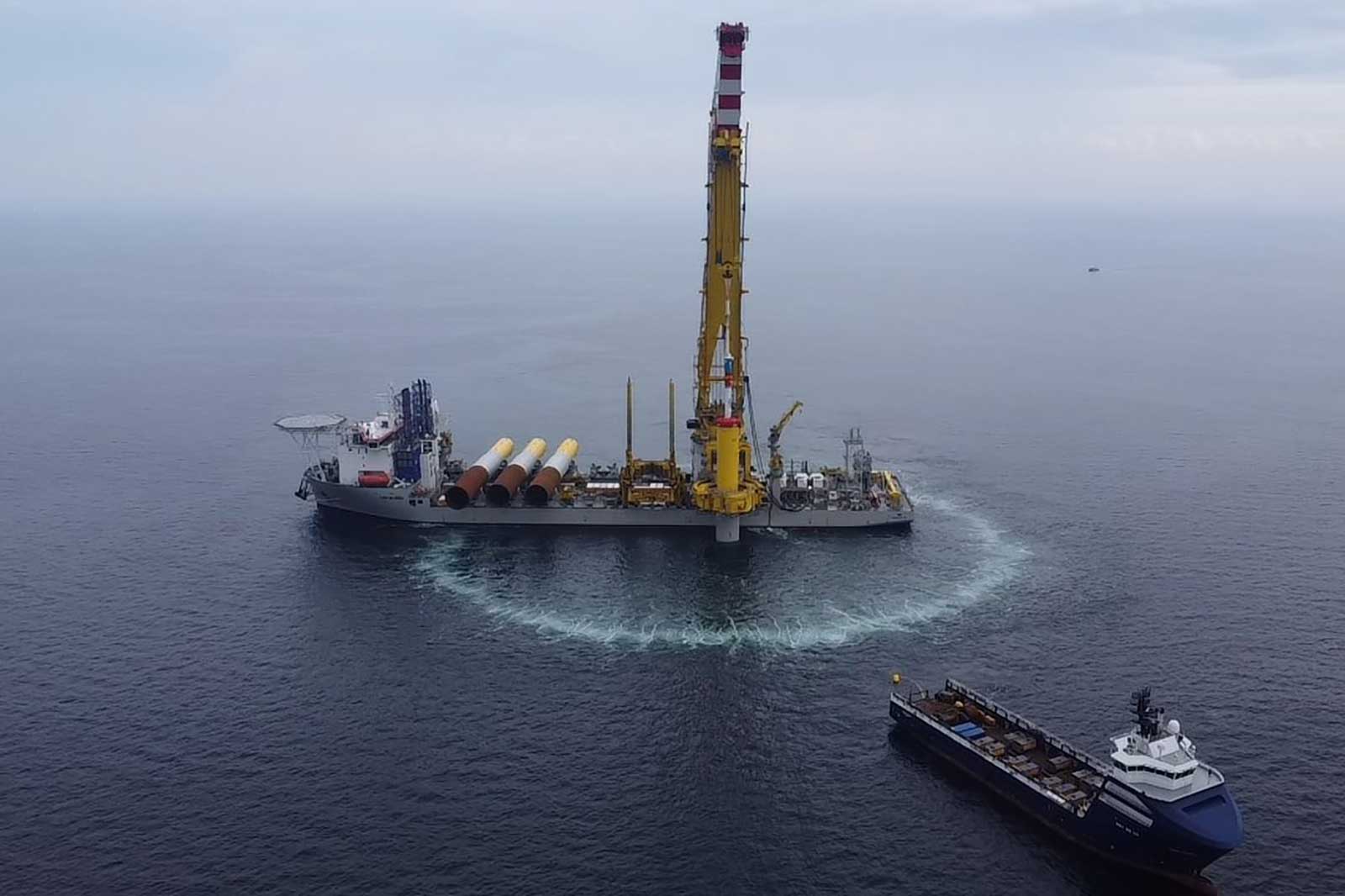 Et skib med en gul kran står i havet, ved siden af et mindre skib med kontainer. Havoverfladen er rolig.