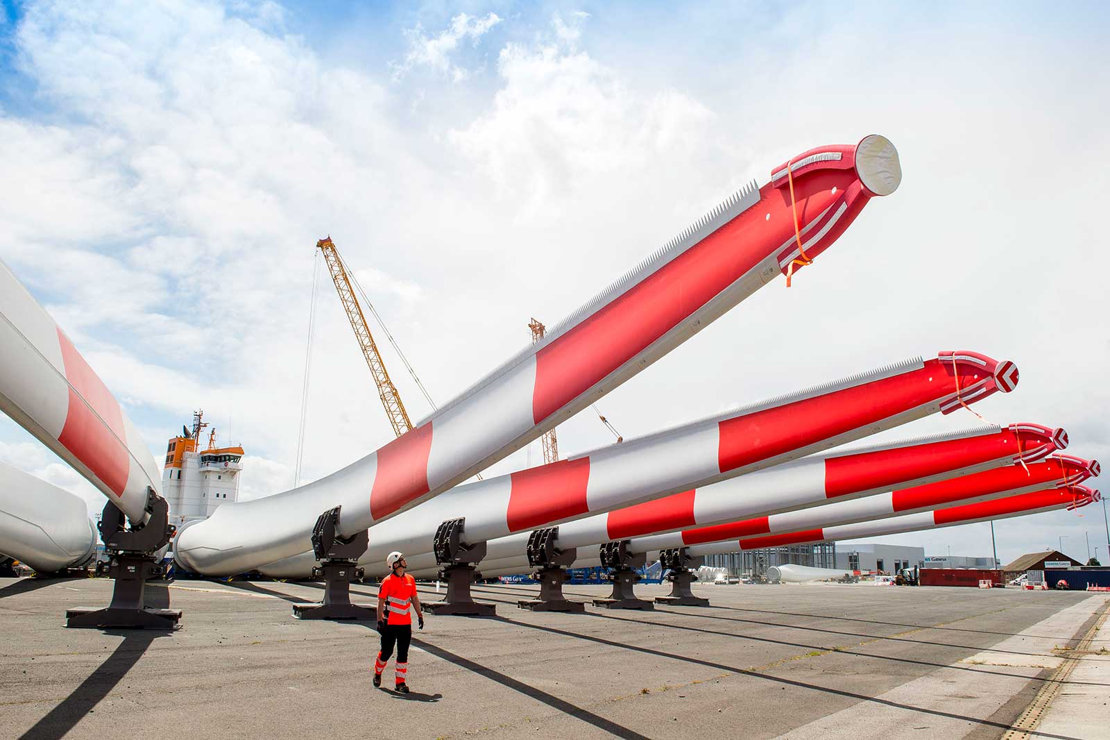 A worker in safety gear walks beside large wind turbine blades with alternating red and white stripes, at a port with cranes.